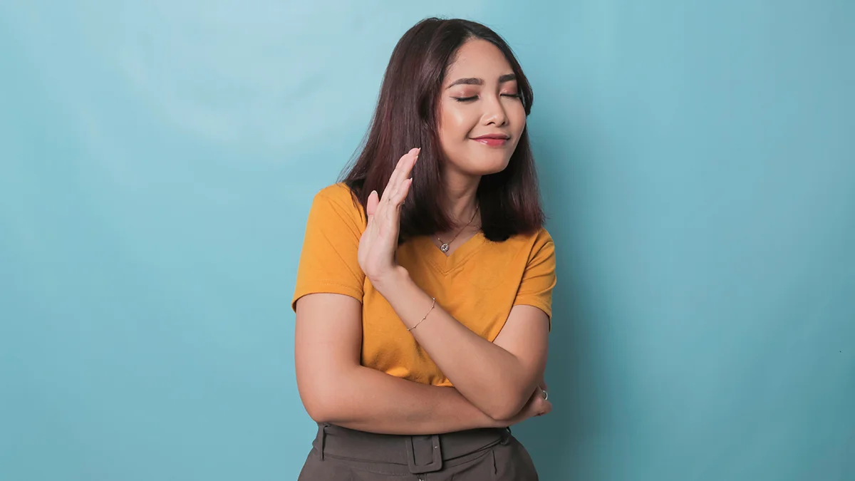 An Asian woman in yellow shirt on blue background politely saying no to something outside of frame